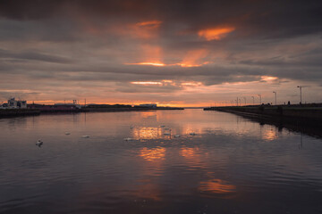 Fototapeta na wymiar Sun rise in Galway city. Dark dramatic sky with orange color sun beams reflects in calm water of river Corrib. Claddagh area. Ireland. Muted colors. Swans colony in the middle of the river