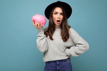 Portrait of shocked amazed dissatisfied young beautiful woman with dark hair wearing stylish gray sweater and black hat isolated over blue background with empty space and holding pink piggy box