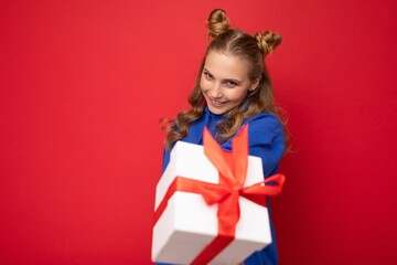 Charming happy funny joyful young blonde woman isolated over red background wall wearing blue trendy hoodie holding gift box and looking at camera