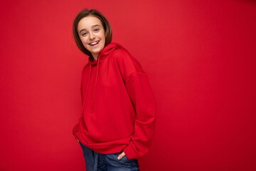Fototapeta premium Shot of beautiful happy cool smiling brunette little girl wearing trendy red hoodie standing isolated over red background wall looking at camera