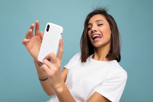 Photo of sexy beautiful smiling happy young brunette woman wearing casual white t-shirt isolated over wall blue background holding and using mobile phone taking selfie looking at gadjet screen and