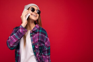 Closeup photo of charming smiling positive young woman wearing hipster purple shirt and casual white t-shirt grey hat and sunglasses isolated over red background holding in hand and talking on mobile