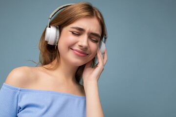 Closeup portrait photo of beautiful positive smiling young blonde woman wearing blue crop top isolated over blue background wall wearing white wireless bluetooth headphones listening to good music and