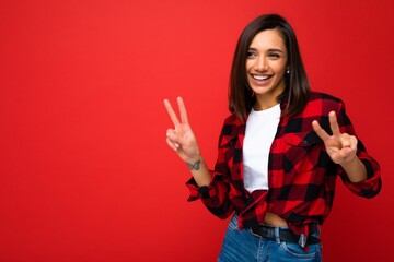 Young happy positive cool beautiful brunette woman with sincere emotions wearing white t-shirt and stylish red check shirt isolated over red background with copy space and showing peace sign