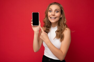 Naklejka premium Photo of beautiful smiling young woman good looking wearing casual stylish outfit standing isolated on background with copy space holding smartphone showing phone in hand with empty screen display for