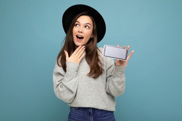 Shot of thoughtful charming young happy woman wearing black hat and grey sweater thinking about something having an idea holding phone looking to the side isolated on background