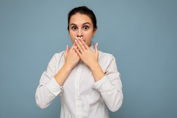 Portrait of young amazed shocked beautiful brunet female person with sincere emotions wearing white shirt isolated over blue background with copy space and covering mouth with hands
