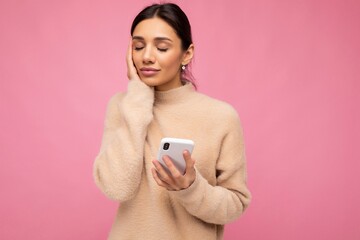 Attractive cute young brunet woman wearing beige warm sweater standing isolated over pink background surfing on the internet via phone and thinking about something pleasant