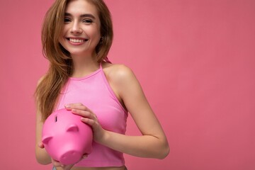 Portrait photo of happy positive smiling young beautiful attractive blonde woman with sincere emotions wearing pink summer top isolated over pink background with copy space and holding pink piggy box