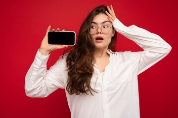 Photo of beautiful shocked young woman good looking wearing casual stylish outfit standing isolated on background with copy space holding smartphone showing phone in hand with empty screen display for
