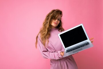 Naklejka premium Photo of beautiful blond curly woman holding computer laptop with empty monitor screen with mock up and copy space wearing pink sweater isolated over pink wall background