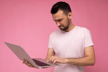 Handsome indignant and disturbing brunet man holding laptop computer and typing on the keyboard looking at netbook monitor in t-shirt on isolated pink background