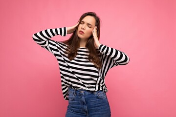 Photo of young sad upset tired attractive brunette female person with sincere emotions wearing casual striped pullover isolated on pink background with copy space and having headache