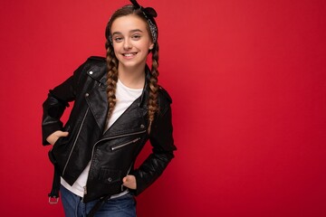 Photo shot of beautiful happy smiling brunette little girl with pigtails wearing trendy black leather jacket and white t-shirt standing isolated over red background wall looking at camera