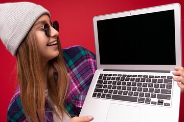 Close-up Portrait of beautiful laughing funny smiling blond young woman holding computer laptop with empty monitor screen wearing sun glasses hat and colourful shirt looking at netbook display and