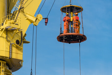 A group of workers consist of riggers and engineers riding a personal transfer basket, transferring...