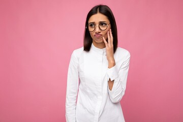 Photo shot of attractive upset sad dissatisfied young brunette female person wearing white shirt and stylish optical glasses isolated over pink background looking to the side
