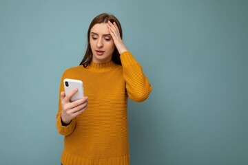 Shocked attractive positive young woman wearing orange sweater poising isolated on blue background with empty space holding in hand and showing mobile phone with empty space for mockup looking at
