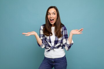 Shot of young positive delightful surprised beautiful brunette woman with sincere emotions wearing trendy check shirt standing isolated on blue background with free space