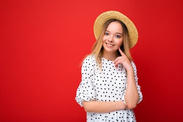 Portrait of young beautiful smiling hipster blonde woman in trendy summer dress and straw hat. Sexy carefree female person posing isolated near red wall in studio. Positive model with natural makeup