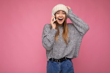 Beautiful positive happy amazed young blonde woman wearing casual grey sweater and beige hat isolated over pink background holding in hand and talking on mobile phone looking at camera