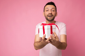 Photo shot of handsome positive happy kind brunette young unshaven man with beard isolated over pink background wall wearing casual pink t-shirt holding white gift box with red ribbon and looking at