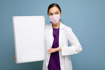 Beautiful brunette female nurse in protective face mask and white medical coat holding an empty magnetic board isolated on blue background