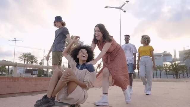 Group of young people having fun with skateboard in the city. Smiling black girl sitting on skateboard being pushing and pulled by caucasian girl. girls.