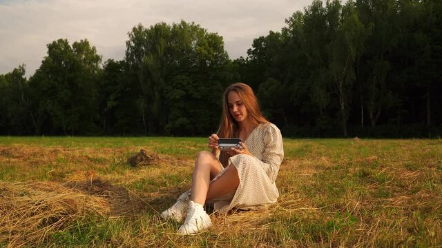 Woman long blond hair, white sundress, sits ground, setting sun field, trees background. Examines, sets up an old retro silver film camera. Preparing photography. An interesting activity, useful hobby