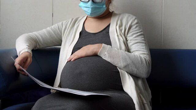 Young Pregnant Woman In Medicine Protective Mask Reading On Couch Waiting For Appointment Medical Control On Hospital. Visit Gynecologist Doctor At Clinic. Examine Expectant Belly Healthcare Check Up