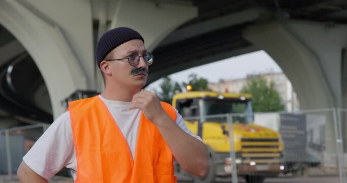 Funny builder or foreman with glasses and orange uniform adjusts mustache AND shakes off hands against car under overpass bridge on street