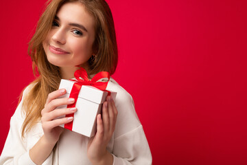 Fototapeta premium Beautiful happy young blonde woman isolated over colourful background wall wearing stylish casual clothes holding gift box and looking at camera