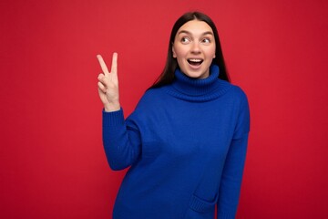 Fototapeta premium Photo of young positive happy smiling beautiful woman with sincere emotions wearing stylish clothes isolated over background with copy space and showing peace gesture with two fingers