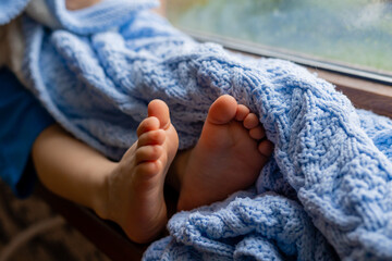 closeup of the legs of a sitting child with bare feet covered with a blue knitted blanket