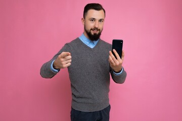 Good looking happy cool young brunette unshaven man with beardwearing stylish grey sweater and blue shirt standing isolated over pink background wall holding smartphone and using phone looking at