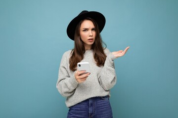 Beautiful young brunette woman thinking wearing black hat and grey sweater holding smartphone looking to the side texting isolated on background