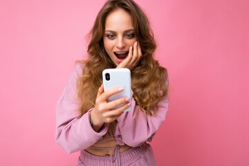 Photo of attractive crazy amazed surprised young woman wearing casual stylish clothes standing isolated over background with copy space holding and using mobile phone looking at telephone screen