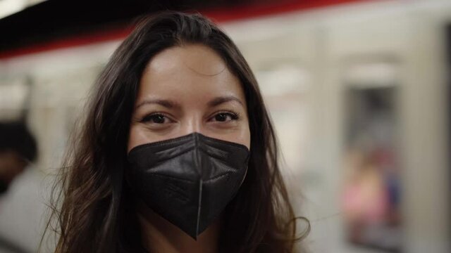 Young woman with mask, looking at camera. Blurred subway train moving in background.