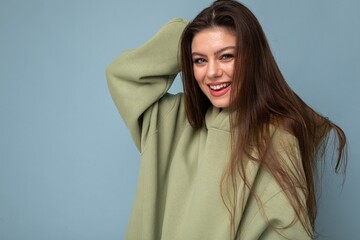 Portrait of young beautiful smiling girl in stylish hipster green hoodie. Sexy carefree woman posing near blue wall. Positive model having fun