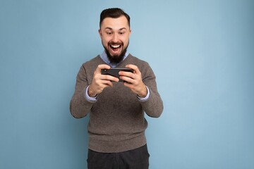 Shot of happy amazing shocked handsome young brunette unshaven man with beard wearing everyday grey sweater and blue shirt isolated on background wall holding smartphone playing games via mobile phone