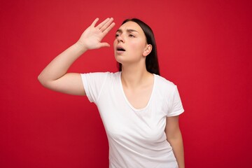 Portrait of emotional young beautiful brunette woman with sincere emotions wearing white t-shirt for mockup isolated over red background with free space and looking to the side with hand