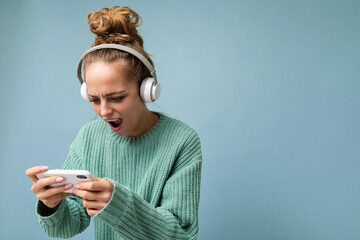Pretty emotional positive young woman wearing blue sweater isolated over blue background wearing white bluetooth wireless headphones and listening to music and using mobile phone watching videos