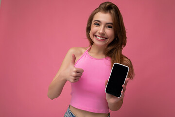 Beautiful joyful young blonde woman wearing pink top poising isolated on pink background with empty space holding in hand and showing mobile phone with empty display for mockup looking at camera and