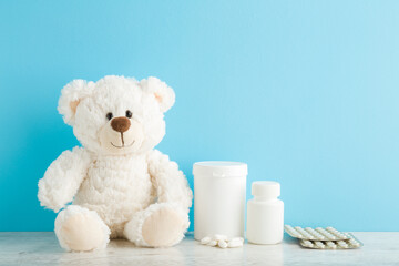 White teddy bear, plastic bottles and blister packs with pills on table at light blue wall...