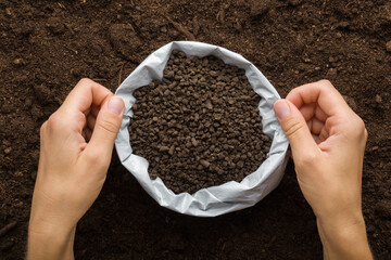 Young adult woman hands holding opened plastic bag with black granules of chicken manure on dark soil background. Closeup. Product for root feeding of vegetables, flowers and plants. Top down view.