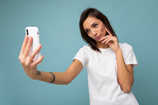 Photo of beautiful smiling happy young brunette woman wearing casual white t-shirt isolated over wall blue background holding and using mobile phone taking selfie looking at gadjet screen