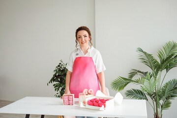 Confectioner chef wearing pink apron posing next to delicious desserts on the white table.