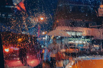 View of rain on car window in focus, during the rainy night, out of focus through the glass is a woman carrying handbag and shopping bags with an umbrella, with streetlights and street traffic