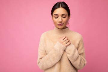 Portrait of young positive cute pretty brunette woman with sincere emotions wearing beige sweater isolated over pink background with empty space and holding hand on chest with closed eyes