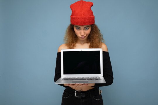 Beautiful funny amusing thoughtful young dark blond curly woman wearing black crop top and red and orange do-rag isolated over light blue wall background holding computer laptop with empty screen